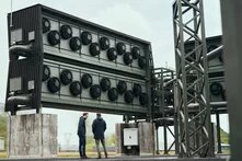 Direct Air Capture in Iceland Zwei Personen stehen vor einer großen, industriellen Anlage mit mehreren Lüftern. Stahlträger und Rohre sind sichtbar.
