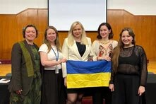 Foto: Fünf Personen stehen in einem Saal vor einer Leinwand und halten gemeinsam eine ukrainische Flagge.