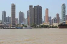 Belém, Brazil Several high-rise buildings behind the riverbank with a boat and houses in the foreground, smoke rising between the trees.