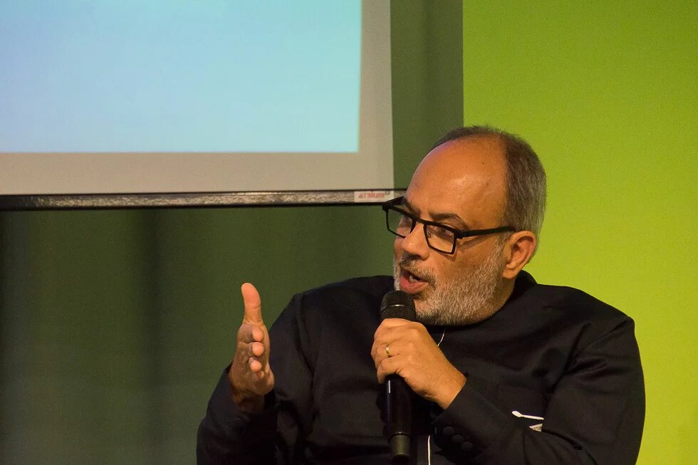 Carlos Lopes A man with glasses speaks into a microphone and makes gestures with his left hand; behind him is part of a screen.
