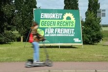 Wahlplakat der Grünen mit dem Schriftzug Gegen Rechts im Mittelpunkt. Im Vordergrund fährt eine Person auf einem Roller vorbei.