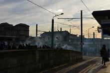 GPT  Foto: Eine Menschenmenge steht auf einem Platz vor dem Bahnhof Saint-Denis in Paris, umgeben von Straßenbahnschienen und Oberleitungen. Rauch steigt auf.