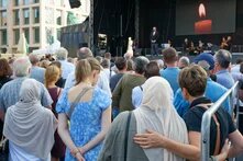 Foto: Menschenmenge bei der Andacht auf dem Neumarkt in Solingen, einige mit gesenkten Köpfen. Auf einer Bühne spricht eine Person, im Hintergrund ist ein Bild einer Kerze.