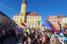 Foto: Menschenmenge demonstriert vor dem Rathaus einer Stadt mit gelber Fassade. Viele halten Regenbogen- und EU-Fahnen. Schilder und Ballons sind sichtbar.