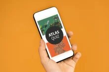 Aufmacherbild - Hintergrund orange, darauf eine Handy haltende Hand. Im Handy zwi Atlanten und ein schwarzer runder Button mit weißer Aufschrift "Atlas-Quiz" 