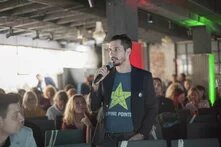 Foto: Ein Mann spricht mit Mikrofon in einer Konferenz. Im Hintergrund sitzen mehrere Menschen und hören zu. Der Raum hat eine industrielle Decke.