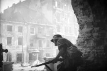 Foto: Soldat in Deckung mit Gewehr, umgeben von Ruinen einer zerstörten Stadt im Hintergrund.