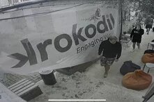 Drei Personen gehen durch einen Außenbereich mit Sitzsäcken. Ein großes Banner mit der Aufschrift „Krokodil“ hängt an der Wand.