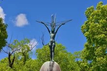 Friedensdenkmal für Kinder in Hiroshima, Japan Foto: Statue eines Kindes mit ausgestreckten Armen, das einen Kranich hält. Umgeben von Bäumen, blauer Himmel im Hintergrund.