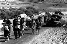 GPT Icon  Photo: A group of people, including children, walk along a road carrying belongings. A destroyed truck and more people are in the background.