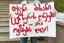 "Ihr Ziel ist es, Zensur durchzusetzen, nicht, Familien zu schützen." Foto: Person steht vor Steintreppe und hält Schild mit georgischer Aufschrift in roter Farbe.