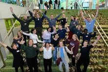 Gruppe von etwa 25 Personen steht auf Treppe, blickt zur Kamera und streckt fröhlich die Arme hoch.