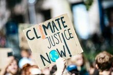 „Climate Justice Now!“ (Klimagerechtigkeit jetzt!) hbs Bildbeschreiber sagte:  Pappschild mit Aufschrift „Climate Justice Now!“ (Klimagerechtigkeit jetzt!) wird von Mensch in Menschenmenge hochgehalten.