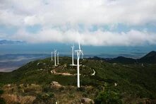 Windräder in Guangdong Sheng, China Auf einem grünen Hügel stehen mehrere Windräder, dahinter das Meer und ein bewölkter Himmel. Ein Weg schlängelt sich durch das Gelände.