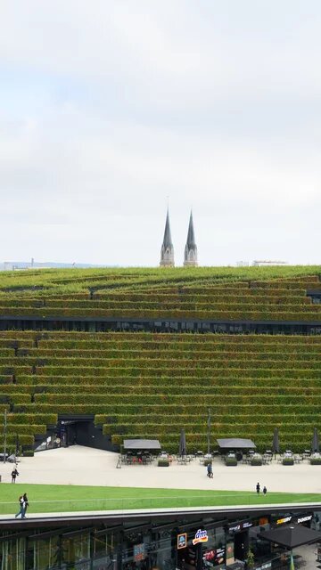 Gebäude mit terrassenförmig bepflanzter Fassade. Umgeben von städtischem Platz, dahinter moderne Gebäude und Kirchtürme.