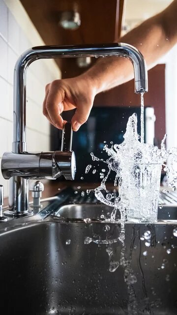 Eine Hand dreht einen Wasserhahn auf, Wasser spritzt in ein überlaufendes Glas in einer Spüle.
