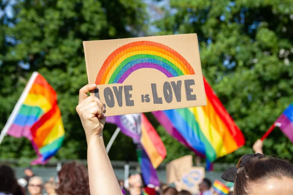 Frauen, die ein Plakatzeichen halten Love ist Liebe mit Regenbogen Symbol der LGBT-Gemeinschaft. Menschenmenge mit Flaggen im Hintergrund. Pride Parade, Gleichstellungsmarsch zur Unterstützung und Feier der LGBT+, LGBTQ Gay lesbische Gleichheit.