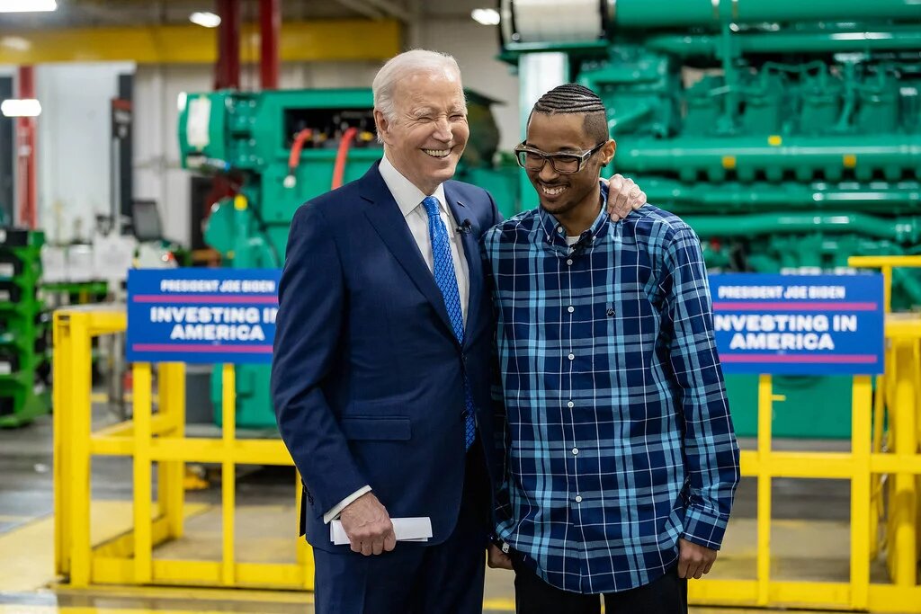 Joe Biden legt seinen Arm um Ausbildungskoordinator Brandon Moore. Im Hintergrund Plakate mit der Aufschrift "INVESTING IN AMERICA"