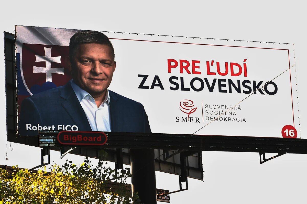 Zu sehen ist ein großes Wahlplakat, auf dem der ehemalige Premierminister Rober Fico abgebildet ist (mittelalt, Seitenscheitel, im Hemd und Jackett, ohne Krawatte)