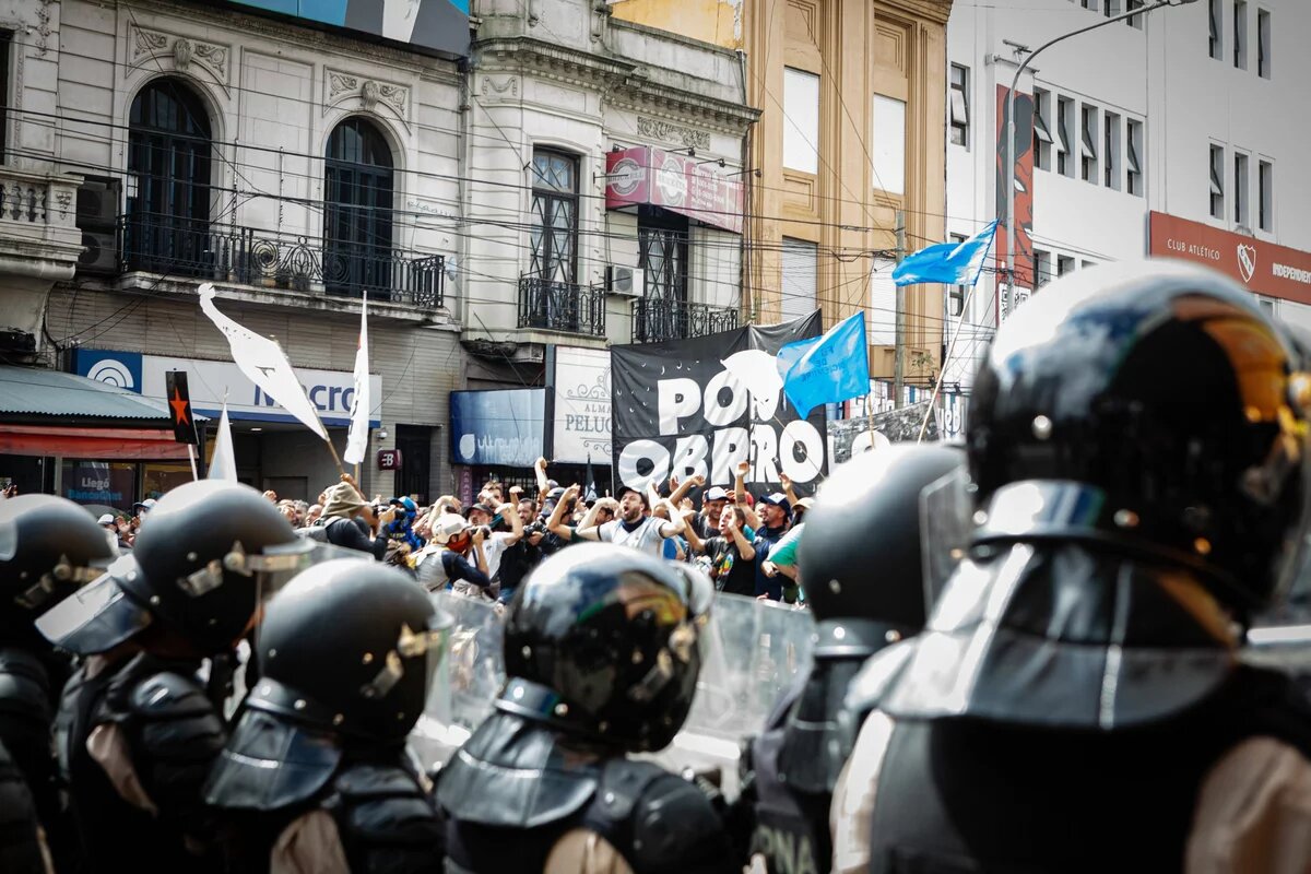 Blick von hinten auf eine Reihe gerüstetet Polizist*innen mit schwarzen Helmen. Ihnen entgegen stehen viele Demonstrierende mit gereckten Fäusten, Fahnen und Schildern.