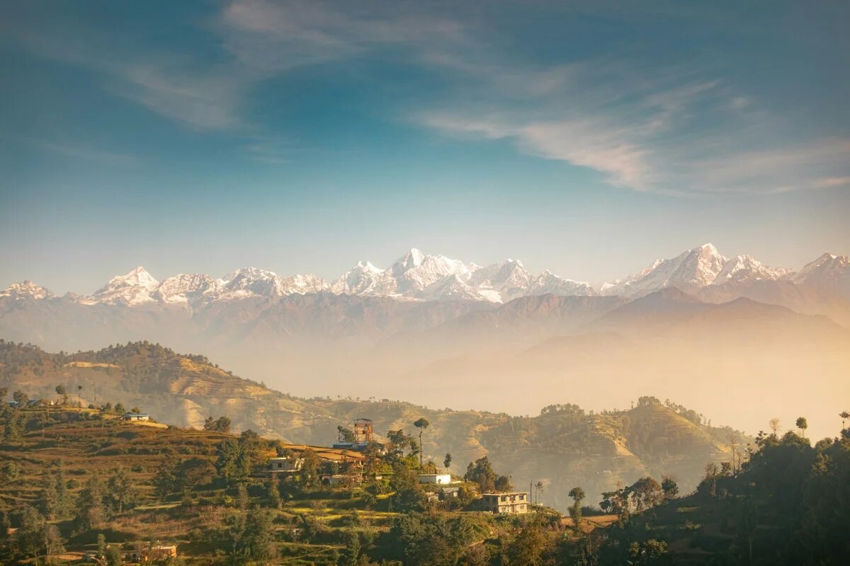 Eine bewaldete Hügelkette mit einigen nepalesischen Häusern, dahinter die schneebedeckten Gipfel des Himalaya unter blauem Himmel.