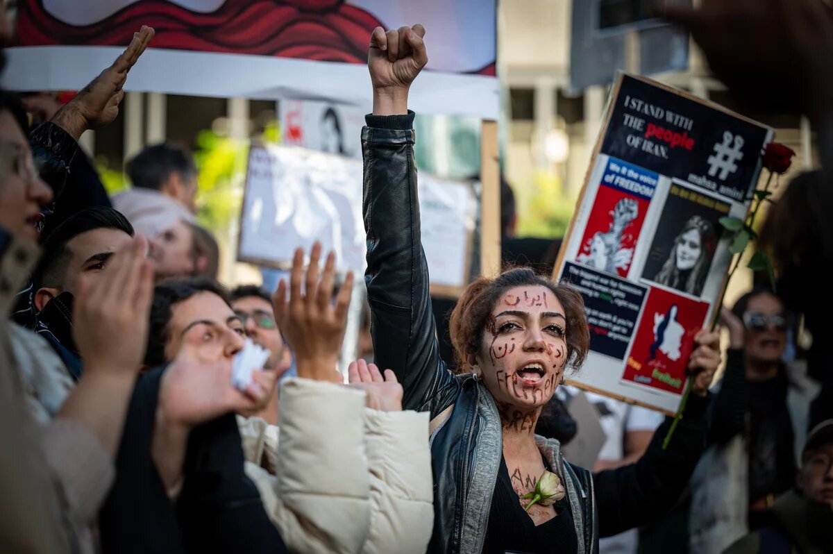 Eine Menschenmenge protestiert in Köln für die Menschenrechte im Iran. Eine Person mit bemaltem Gesicht und erhobener Faust hält ein Schild. Anderen klatschen und halten Schilder hoch.  A crowd protests for human rights in Iran in Cologne | A person with a painted face and a raised fist holds a sign. Others clap and hold up signs.