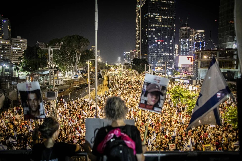 Foto: Nachtaufnahme in Tel Aviv, viele Demonstrant*innen mit Fahnen und Plakaten auf einer Straße, Wolkenkratzer im Hintergrund.
