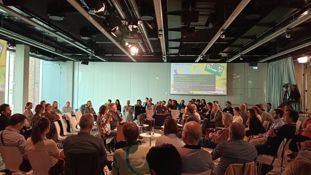 Photo: In a modern conference room, many people sit in a circle of chairs in several rows. Two people are speaking in the middle, with a screen with text in the background