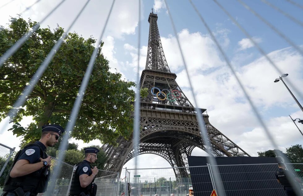 Eiffelturm mit Olympischen Ringen, bewacht von zwei Polizisten links. Bäume im Hintergrund. Absperrzaun im Vordergrund.
