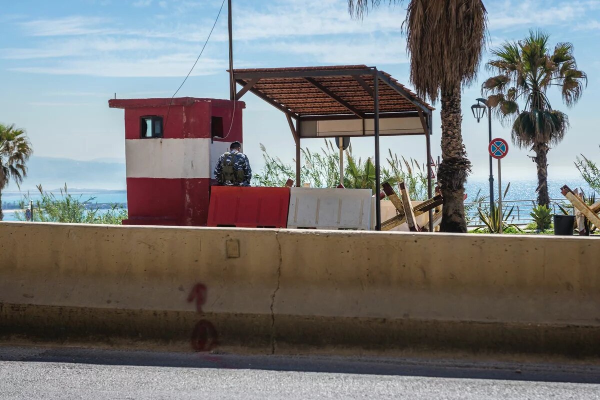 Foto: Ein Kontrollpunkt am Meer, ein uniformierter Mensch sitzt in einem rot-weißen Wachhäuschen, Palmen im Hintergrund.