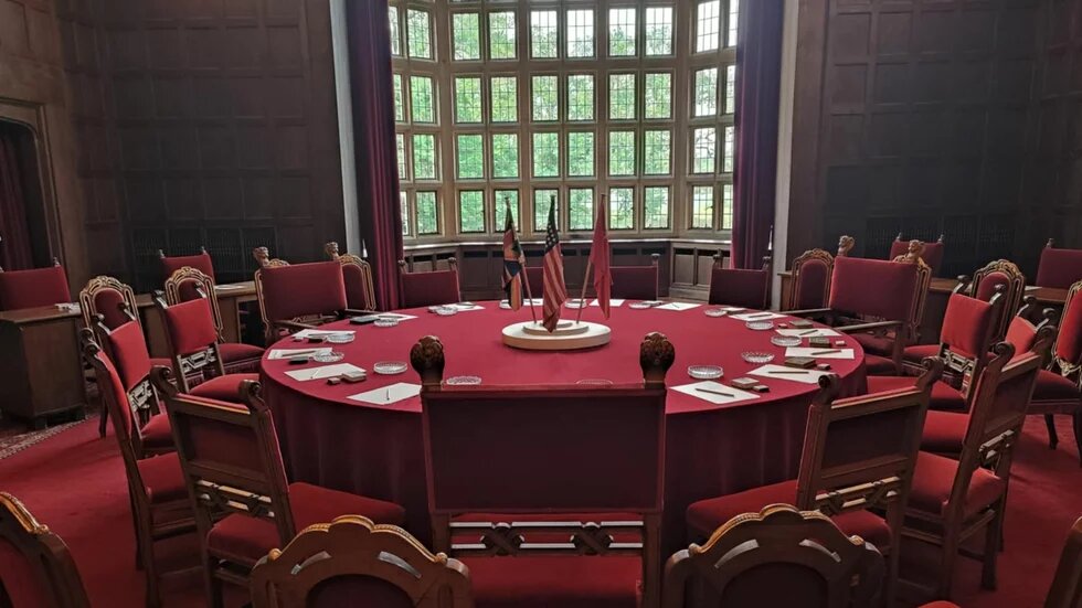 Photo: Conference room in Cecilienhof with a round table, red-upholstered chairs, and large windows in the background. British, American, and Soviet flags in the center of the table.