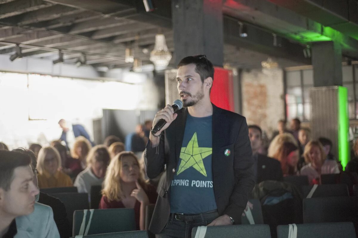 Foto: Ein Mann spricht mit Mikrofon in einer Konferenz. Im Hintergrund sitzen mehrere Menschen und hören zu. Der Raum hat eine industrielle Decke.