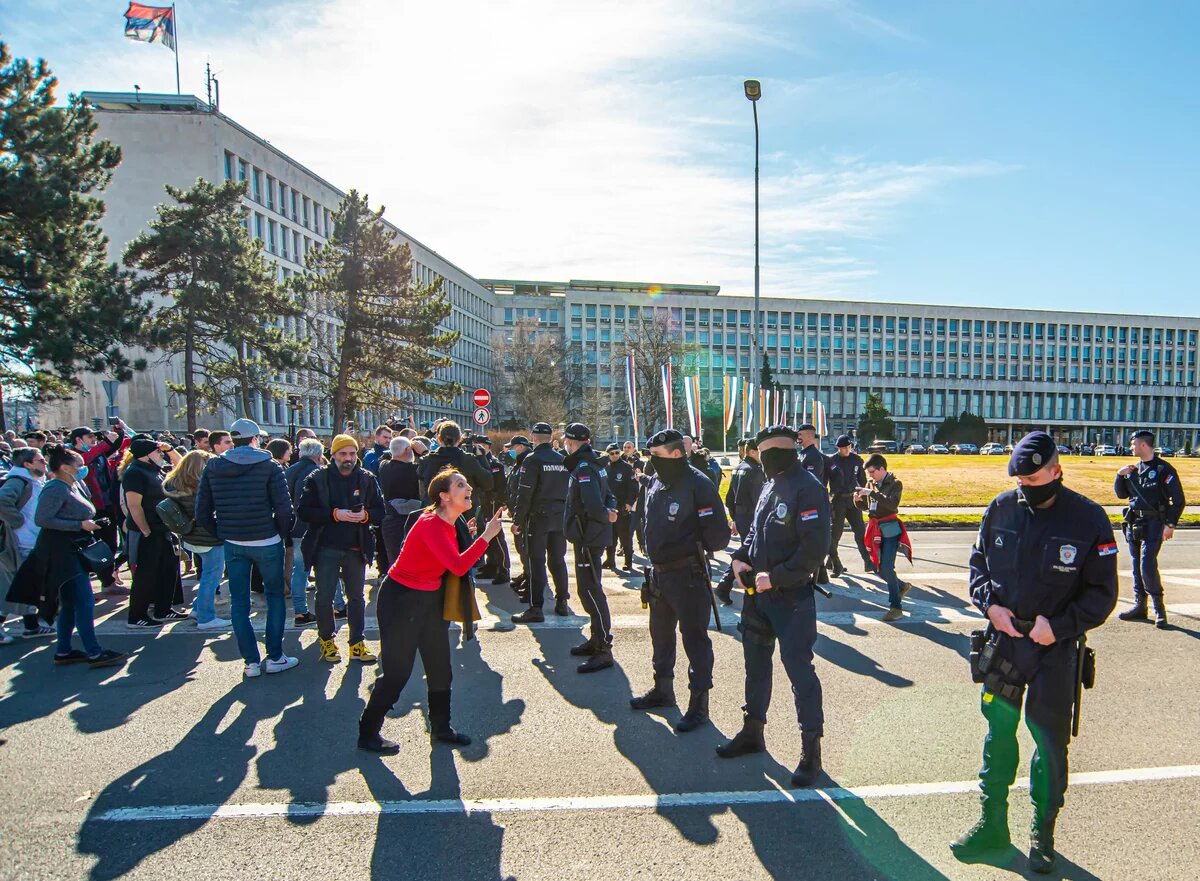 Foto: Eine Gruppe serbischer Polizist*innen steht einer Menge von Demonstrierenden gegenüber. Im Hintergrund ist ein großes, rechteckiges Gebäude.