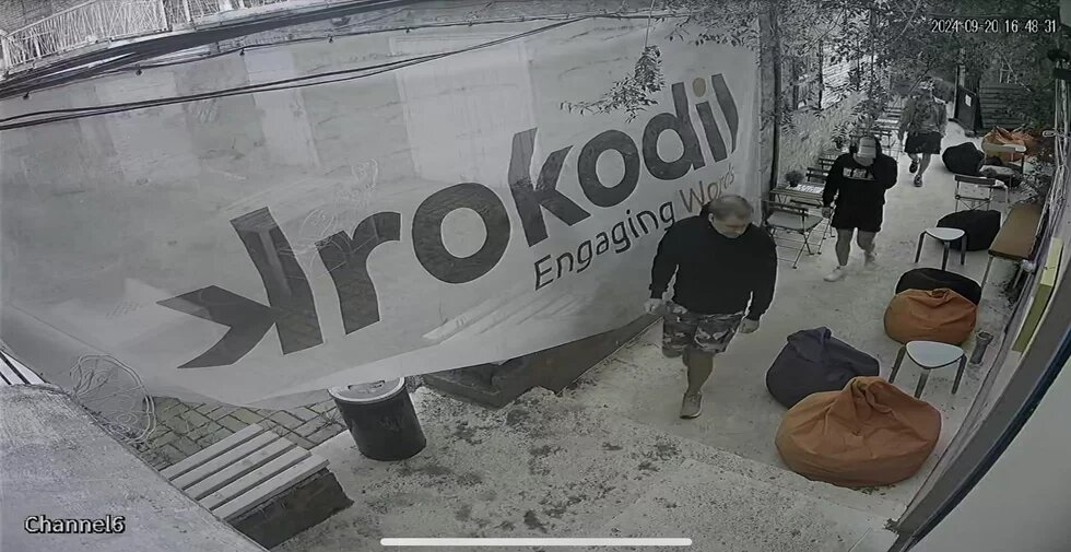 Drei Personen gehen durch einen Außenbereich mit Sitzsäcken. Ein großes Banner mit der Aufschrift „Krokodil“ hängt an der Wand.