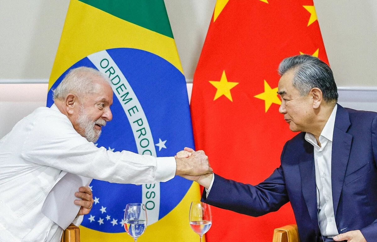 Foto: Luiz Inácio Lula da Silva und Wang Yi geben sich die Hand. Im Hintergrund sind die Flaggen Brasiliens und Chinas.