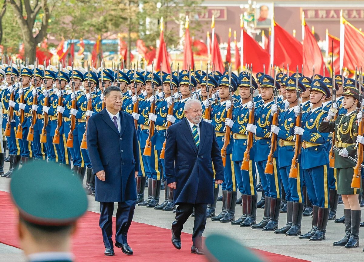 Foto: Xi Jinping und Luiz Inácio Lula da Silva schreiten eine Ehrengarde entlang. Chinesische Soldat*innen stehen in Reihen mit Gewehren und roten Fahnen im Hintergrund.