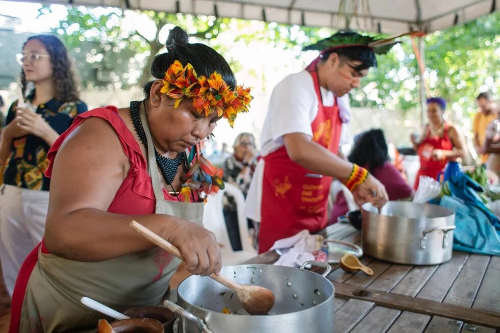 Zubereitung des Essens bei einer Gemeinschaftsmahlzeit. Foto: Zwei Personen mit buntem Kopfschmuck und Schürzen kochen an einem Holztisch im Freien. Weitere Menschen sind im Hintergrund sichtbar.