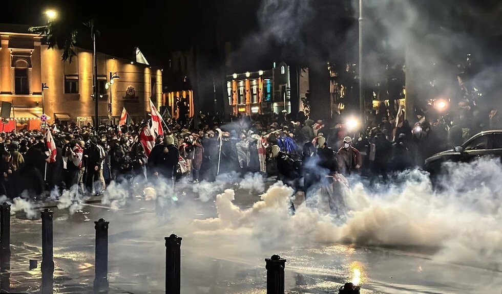 Protest in Georgien, 04.10.2025 - Polizisten setzen während einer Massenkundgebung der Opposition Tränengas gegen Demonstranten ein. Große Menschenmenge bei Nacht, viele Flaggen, Rauch steigt auf, einige Personen tragen Masken, Lichtquellen im Hintergrund.